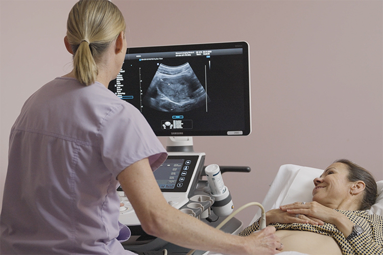 A woman lies on an examination table while a sonographer performs an ultrasound scan using a handheld transducer on her belly.